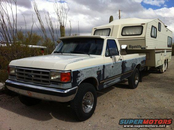 1991 Ford F-250 pickup truck towing an Alpenlite camper, parked on a dirt road with desert vegetation