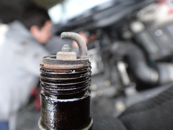 Close-up of a dirty, used spark plug with black deposits and a bent electrode, with an engine and person blurred in the background