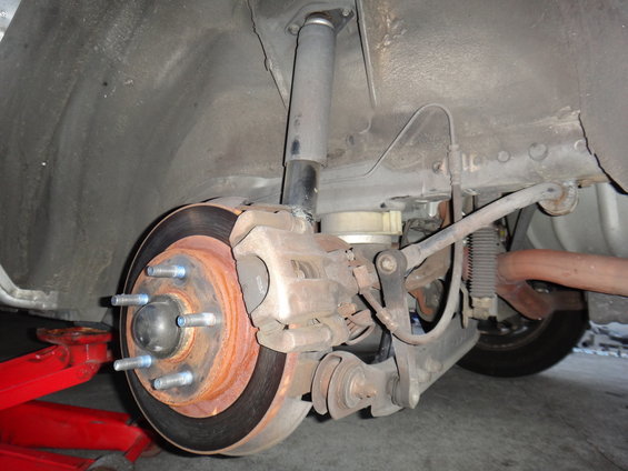 Rear suspension assembly with brake rotor, caliper, and shock absorber visible. Red jack supports the vehicle