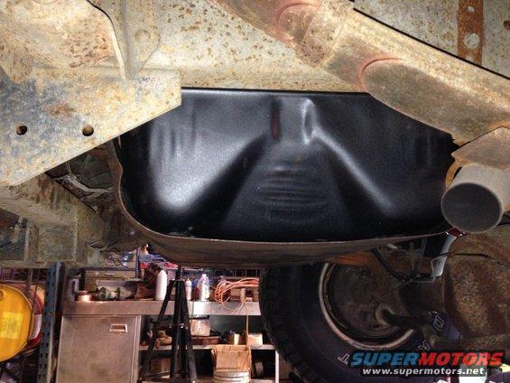Underside view of a vehicle's fuel tank and exhaust pipe, with a workshop in the background