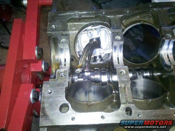 Engine block on red stand, showing a broken connecting rod attached to a piston, with a damaged camshaft