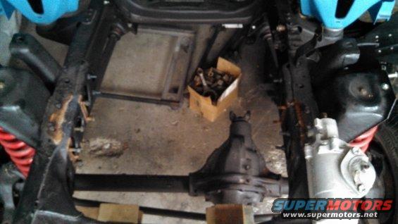 Underside view of a vehicle chassis with suspension and drivetrain parts visible. A box of hardware sits on the floor