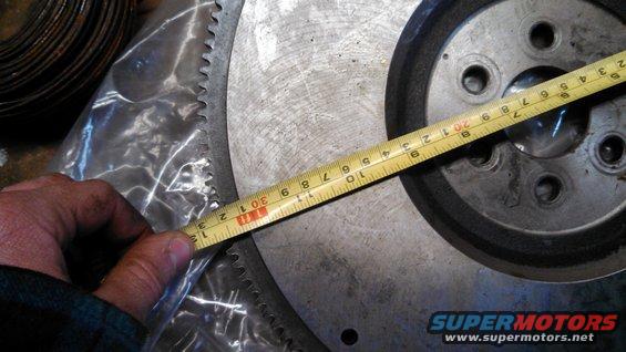 Measuring a flywheel with a tape measure. The flywheel has a toothed outer edge