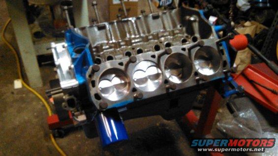 V8 engine block on an engine stand, showing pistons and cylinder bores. Blue engine paint visible