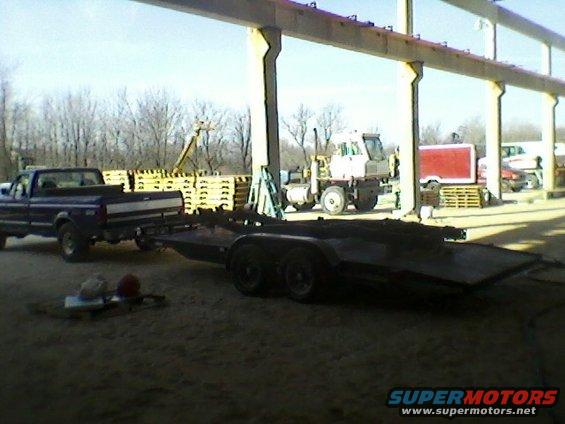 Dark blue pickup truck towing a flatbed trailer, seen from the front-left. The truck appears to be from the early 1990s