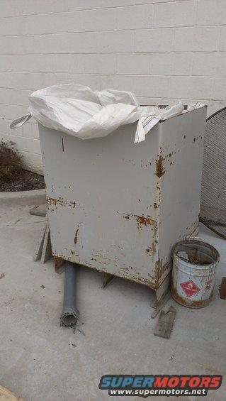 Large, rusty metal container covered by a white bag, with a pipe sticking out the bottom. A bucket with a hazard symbol is nearby