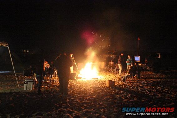 People gathered around a large campfire on a sandy area at night, with some tents and vehicles visible in the background