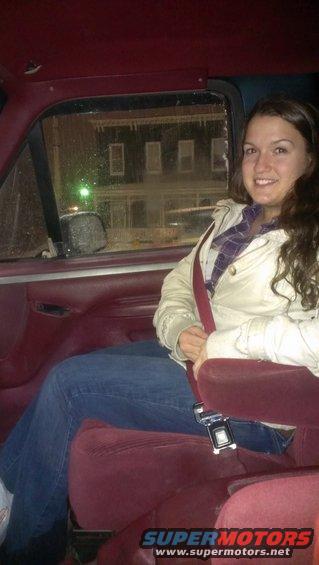 Woman in a maroon interior, wearing a cream jacket and jeans, with a seatbelt fastened