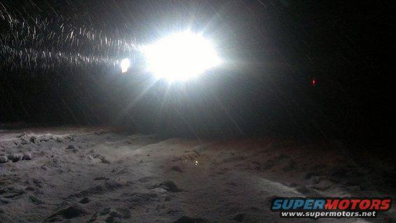 Bright headlight shines through falling snow onto a snowy ground