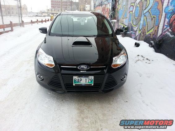 Black 2012 Ford Focus with a hood scoop parked on a snowy street in front of a graffiti-covered wall