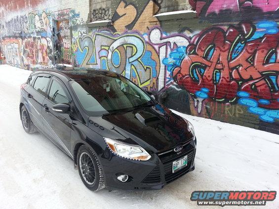 Black hatchback parked on snowy street in front of graffiti-covered wall. License plate is visible