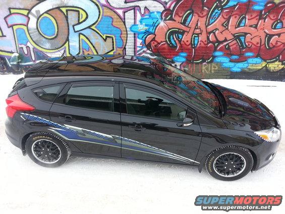 Black 2012 Ford Focus hatchback with blue and silver racing stripes parked in snow, graffiti wall in background