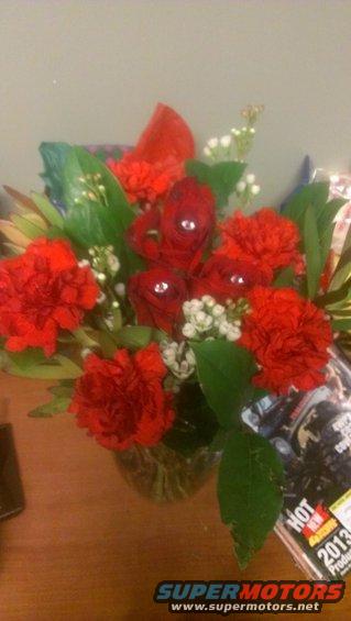 Bouquet of red carnations and roses with white accents in a glass vase on a wooden surface, next to a magazine