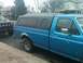 1993 Ford Bronco - A blue pickup truck with a black camper shell is parked next to a black SUV on a residential street.