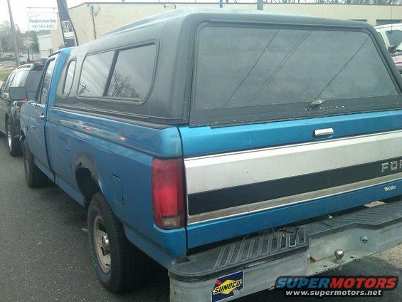 Side view of a blue Ford F-series pickup truck with a black camper shell. Another vehicle is parked to its left