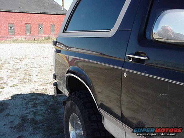 Side view of a 1993 Ford Bronco, showing the rear quarter panel and tire. Red brick building in background
