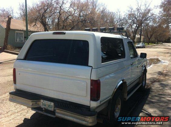 White Ford Bronco with a cap, seen from the rear three-quarter angle. Chrome trim and roof rack with lights are visible