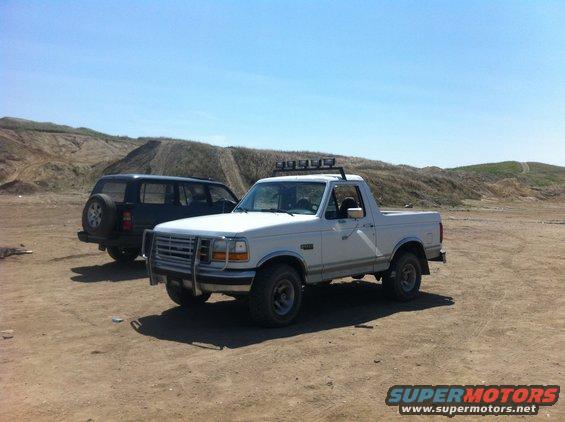 White 1992 Ford Bronco with light bar and brush guard parked next to a black SUV in a dirt lot