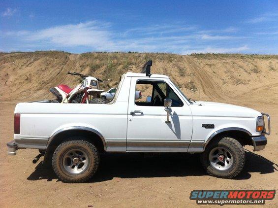White Ford Bronco with a dirt bike in the bed, parked on dirt with a hill in the background