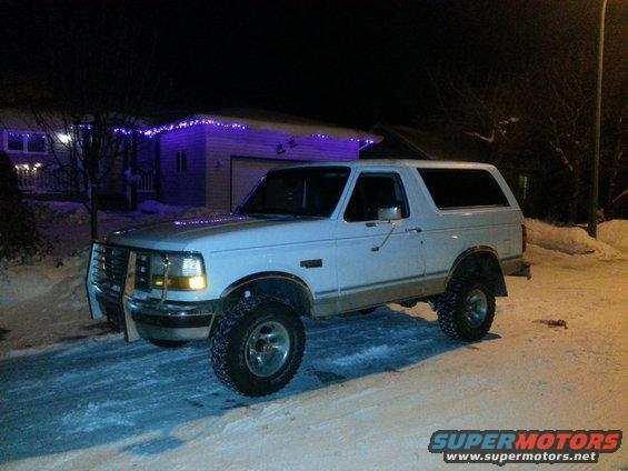 White 1994 Ford Bronco parked on a snowy street at night with purple lights on the house in the background