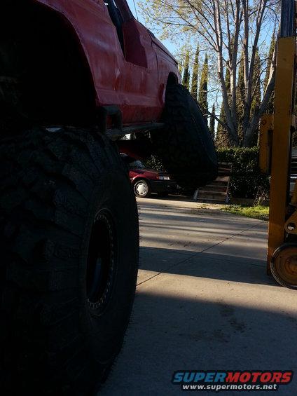 Red off-road vehicle body and massive tire in foreground, forklift to the right, red sedan in background