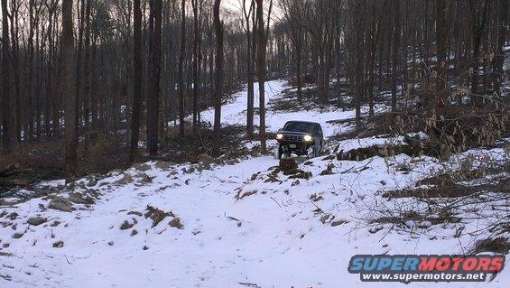 Dark SUV driving on a snowy trail in a forest. The vehicle has a boxy body style