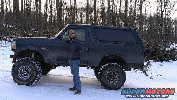 Dark blue Ford Bronco with large tires and a man standing next to it in a snowy wooded area