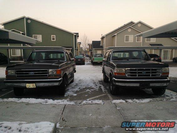 Two Ford Broncos parked in front of apartment buildings. One is dark, the other is dark gray. A green car is in the background