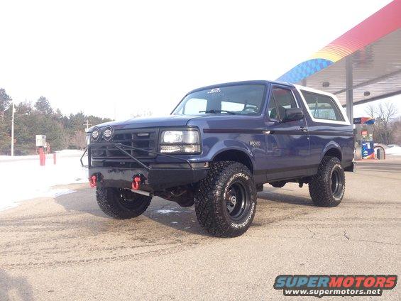 Blue 1996 Ford Bronco with black aftermarket bumper, red tow hooks, and large off-road tires parked at a gas station
