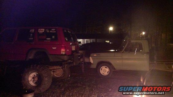 Red SUV with stickers on the side parked next to a green pickup truck at night