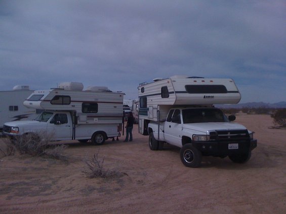 Two white pickup trucks with camper shells parked in a desert landscape under a cloudy sky