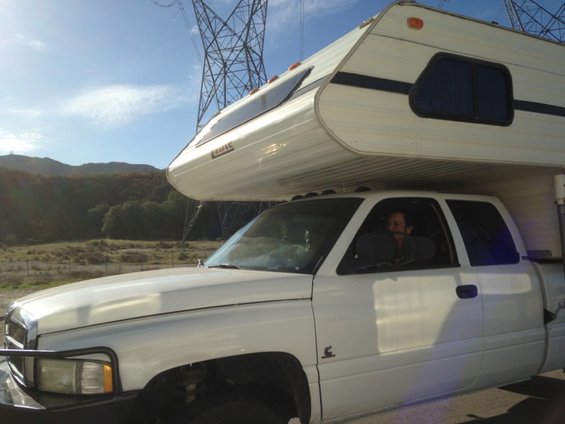 White pickup truck with a white camper shell mounted in the bed. Power lines and hills in background