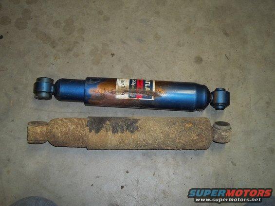 Two shock absorbers on a concrete floor; one is blue with a label, the other is rusty