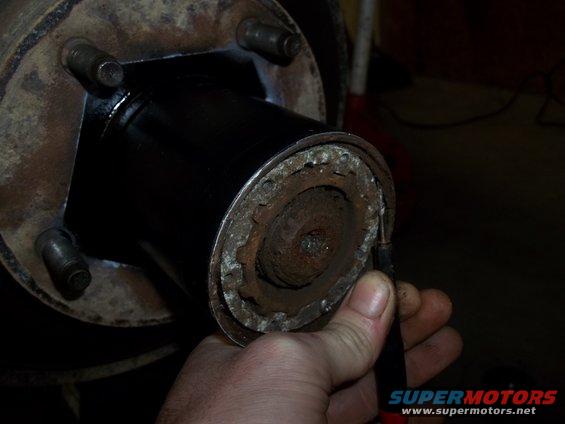 Close-up of a rusty wheel hub assembly with a black painted center section. A hand holds a tool near the edge