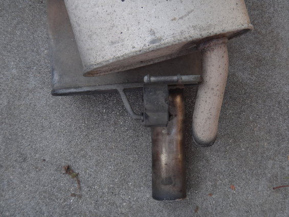 Exhaust muffler and pipe assembly on concrete. The muffler is rectangular and silver, with a curved tailpipe exiting to the right