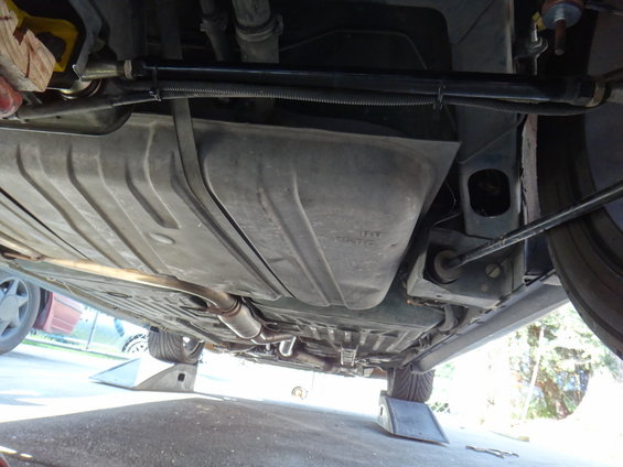 Underside view of a car showing the fuel tank, exhaust pipe, and suspension components. Car is on ramps