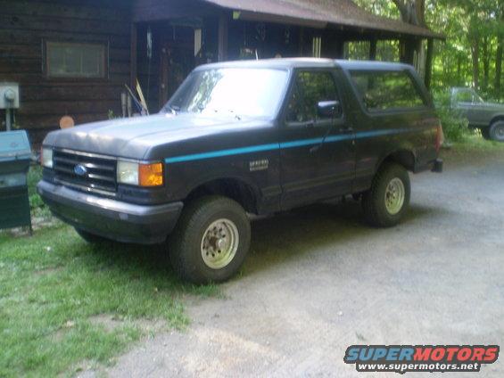 Dark blue Ford Bronco parked in front of a wooden building. Another vehicle is visible in the background