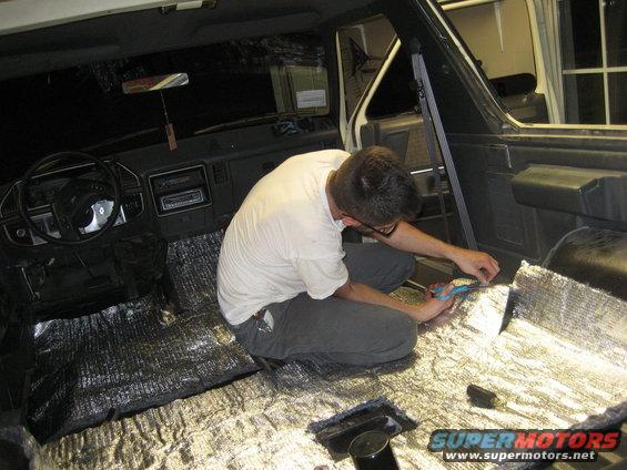 Man cutting silver sound deadening material on the floor of a dark vehicle interior. Steering wheel and dashboard visible