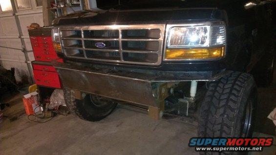 Front of a black Ford truck with a chrome grille and a custom metal bumper. It has large off-road tires and is in a garage