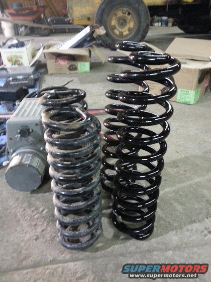 Two coil springs, one used and dirty, the other new and black, sit on a concrete floor next to a gray electrical box