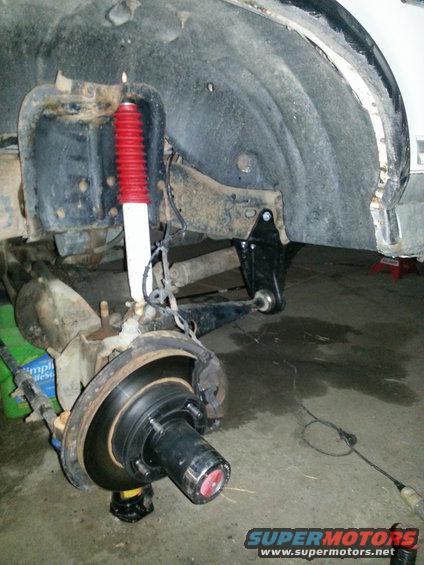 Front suspension assembly of a white vehicle, showing a red and white shock absorber, brake rotor, and hub. The body panel above is rusted