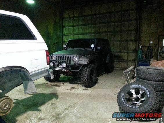 Black Jeep Wrangler parked next to a white SUV in a garage. Tires are stacked nearby