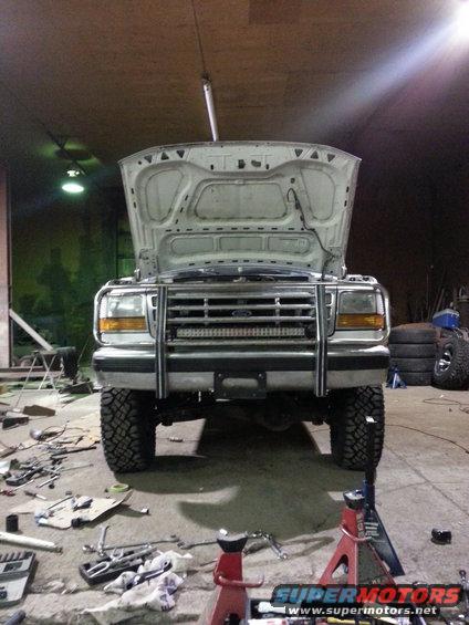 Front view of a white Ford F-Series truck with hood up, bull bar, and LED light bar. Tires are large and knobby. Garage setting with tools on the floor