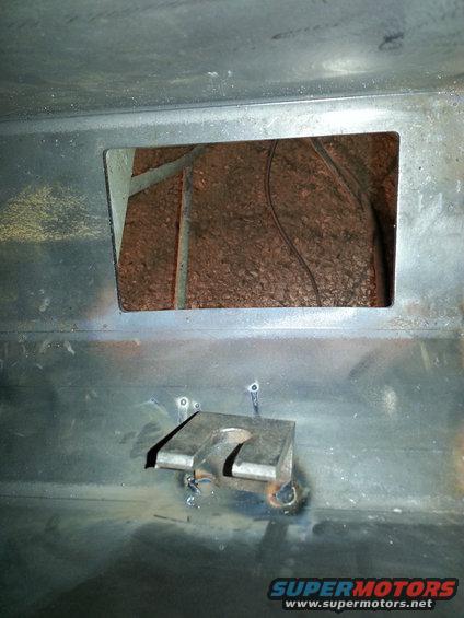 Close-up of a metal bracket welded to a vehicle frame, with a rectangular opening revealing insulation and wires