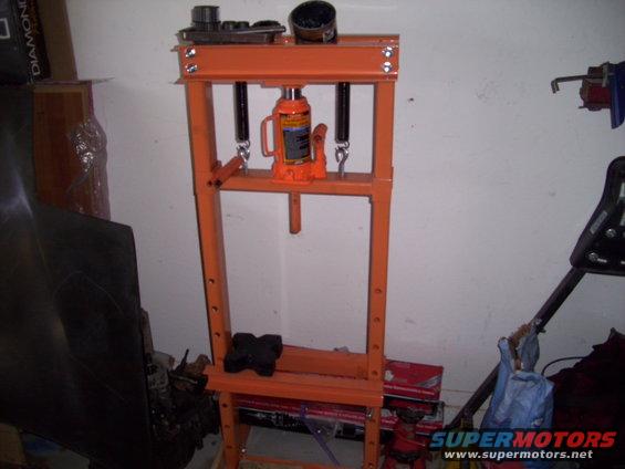 Orange hydraulic press with a bottle jack in a garage setting. Some boxes and car parts are visible