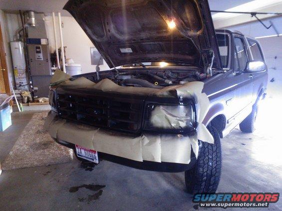 Front of a dark-colored Ford Bronco with hood up, grille and headlights taped off for painting