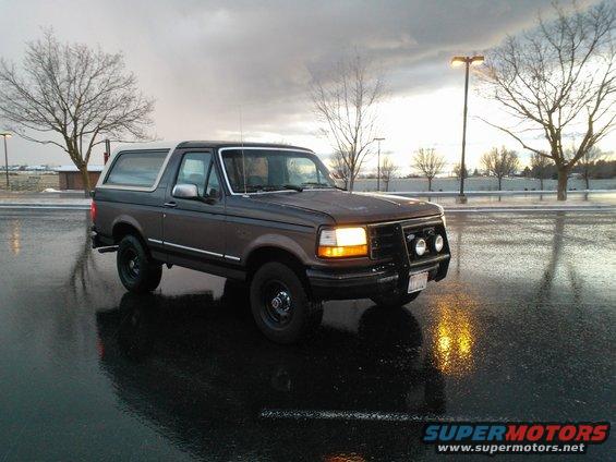 1992 Ford Bronco parked on wet asphalt, front bull bar with lights, overcast sky