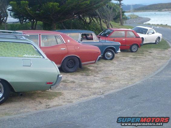 Several cars parked on a dirt lot near water, including a light green station wagon, a red sedan, a blue sedan, a red hatchback, and a white coupe