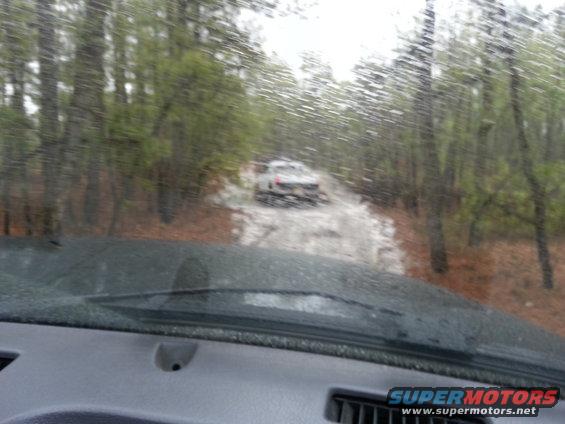 Two white SUVs on a muddy forest road, seen through a rain-streaked windshield