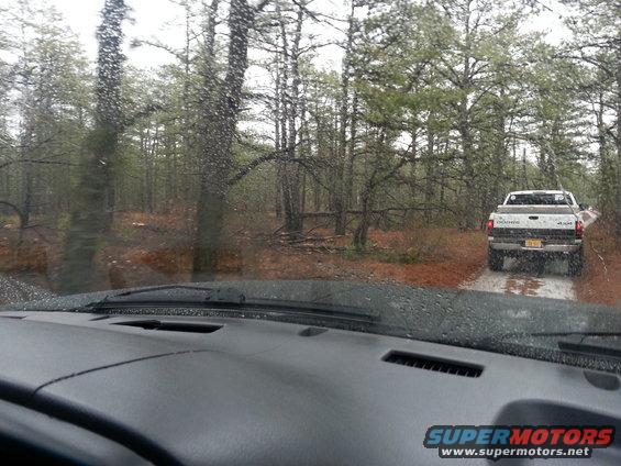 View from inside a vehicle looking at a white Dodge pickup truck driving away on a wet dirt road through a pine forest